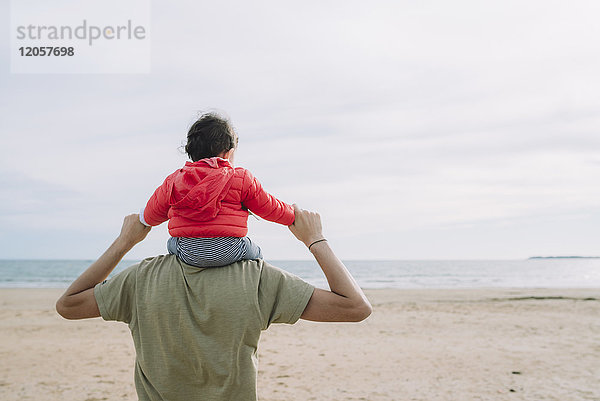 Rückansicht des Vaters  der seine kleine Tochter auf den Schultern am Strand trägt.