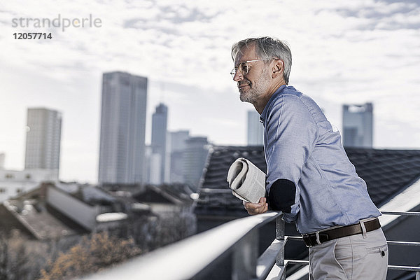 Grauhaariger Mann steht auf dem Balkon mit Zeitungsausschnitt über der Stadt
