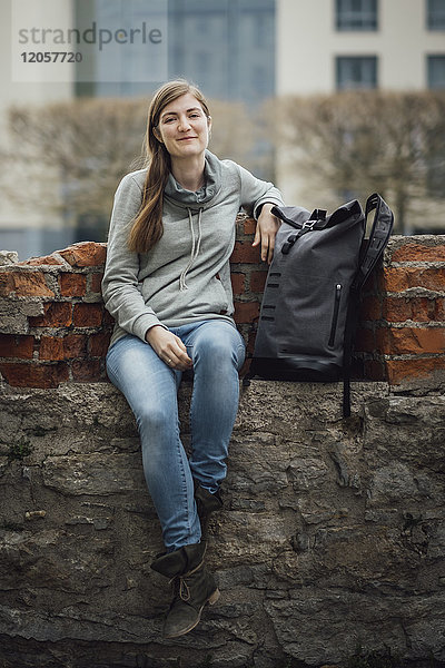 Porträt einer lächelnden jungen Frau  die mit ihrem Rucksack an der Wand sitzt.