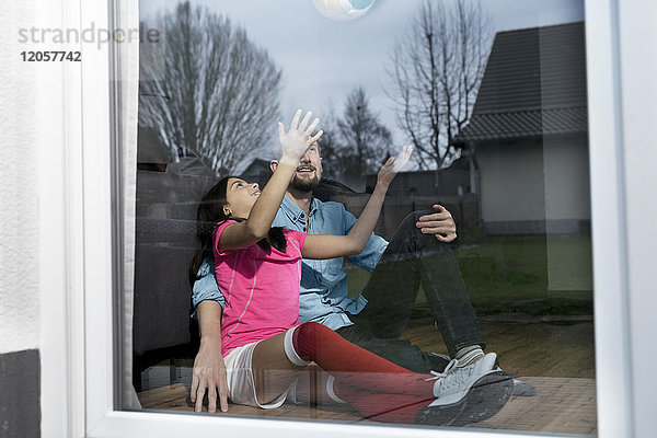 Mädchen im Fußball-Outfit sitzt neben Vater auf dem Boden im Wohnzimmer und spielt mit dem Fußball.