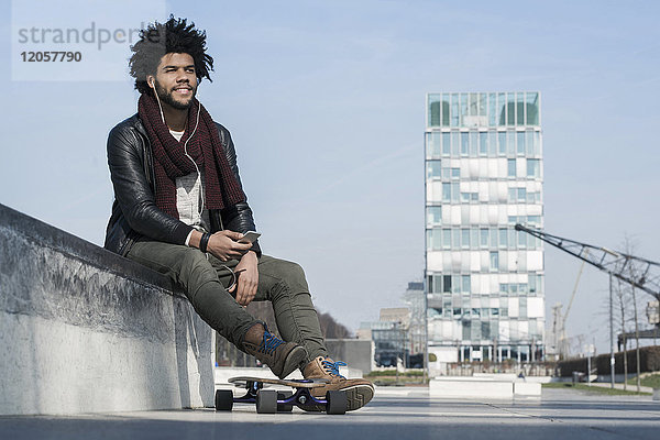 Lächelnder Mann mit Longboard im Skatepark beim Musikhören auf seinem Smartphone
