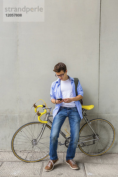 Junger Mann mit Rennrad beim Blick auf das Handy vor der Betonwand