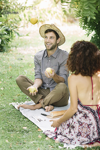 Ein glückliches Paar beim Picknick im Park mit einem Mann  der mit Äpfeln jongliert.