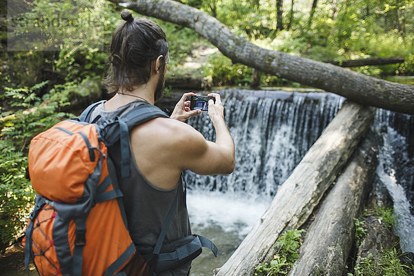 Junger Mann macht ein Handyfoto an einem Wasserfall im Wald