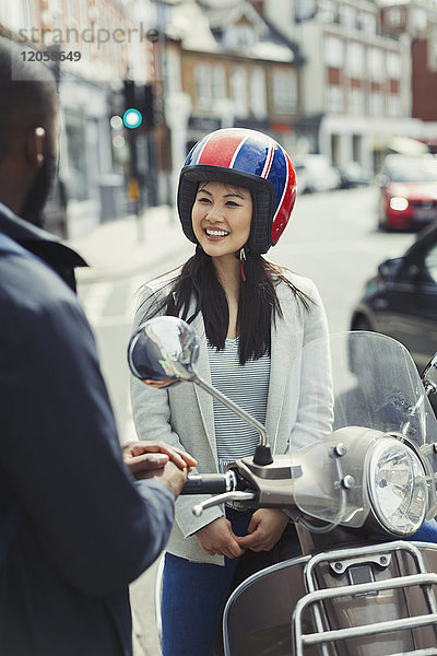 Lächelnde junge Frau im Helm auf dem Motorroller  im Gespräch mit einem Freund in der Stadt.