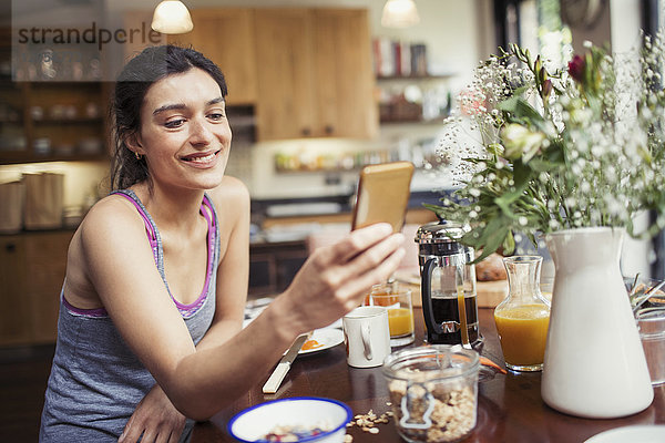 Lächelnde junge Frau SMS mit Smartphone am Frühstückstisch
