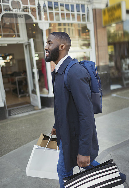 Lächelnder junger Mann  der an der Schaufensterfront entlanggeht und Einkaufstaschen trägt.
