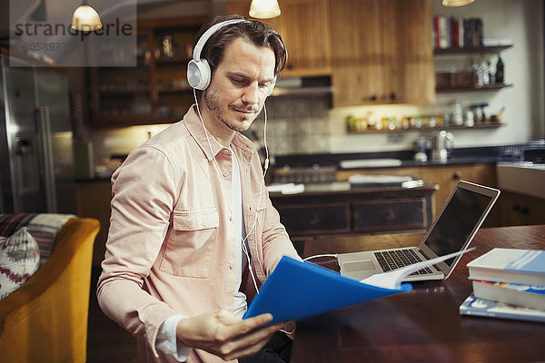 Mann mit Kopfhörern arbeitet am Laptop  liest Papierkram in der Küche