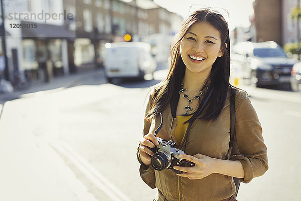 Portrait lächelnde  selbstbewusste junge Touristin mit Kamera auf sonniger Stadtstraße