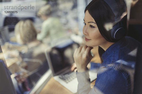 Nachdenkliche junge Frau am Laptop Musik hören mit Kopfhörern am Caféfenster