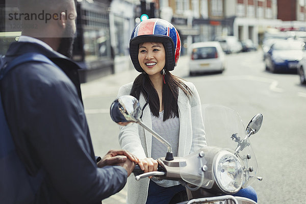 Lächelnde junge Frau auf dem Motorroller im Gespräch mit einem Freund auf einer sonnigen Stadtstraße.