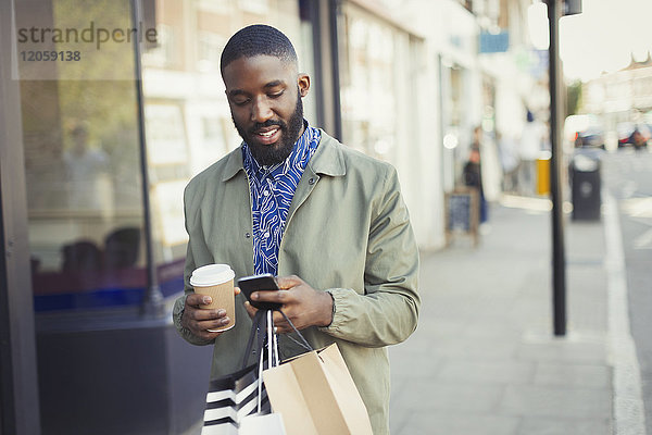Junger Mann mit Kaffee und Einkaufstaschen SMS mit Handy auf städtischem Bürgersteig