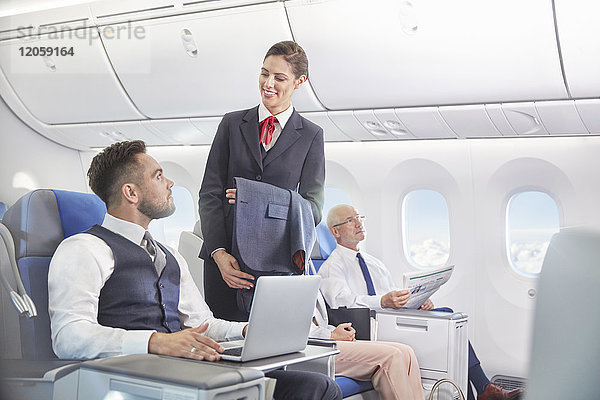 Flugbegleiterin im Gespräch mit Geschäftsmann am Laptop im Flugzeug