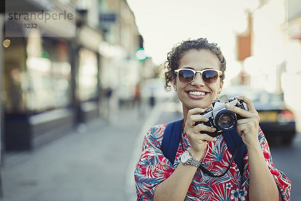 Portrait lächelnde junge Touristin mit Sonnenbrille fotografiert mit Kamera auf urbaner Straße