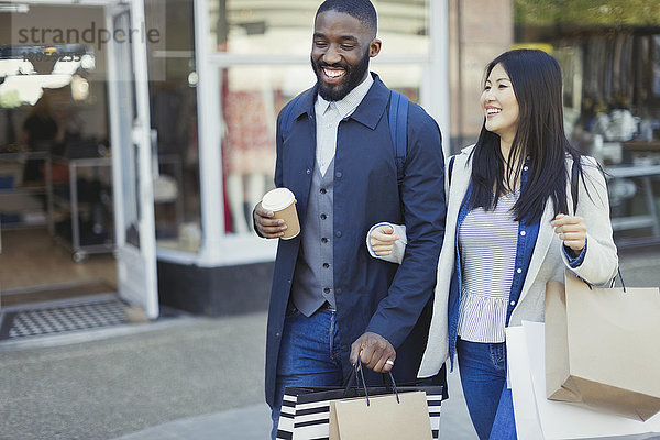 Lächelndes junges Paar geht Arm in Arm entlang der Schaufensterfront mit Kaffee und Einkaufstaschen.