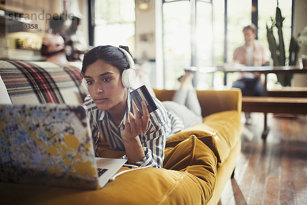 Junge Frau mit Kopfhörer und Kreditkarte online einkaufen am Laptop auf dem Wohnzimmersofa