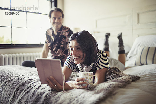 Lächelndes Paar  das sich entspannt  Kaffee trinkt und digitale Tabletten auf dem Bett benutzt.