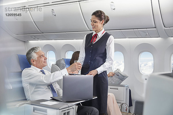 Flugbegleiterin serviert Getränk für Geschäftsmann  der am Laptop im Flugzeug arbeitet