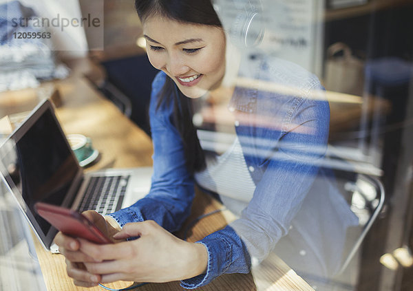 Lächelnde junge Frau beim Musikhören mit Kopfhörer und SMS mit Handy am Laptop im Caféfenster