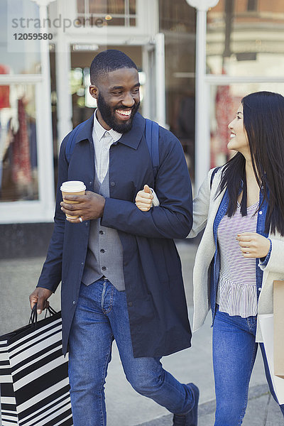 Lächelndes junges Paar  das mit Kaffee und Einkaufstüten an der Fassade entlanggeht.