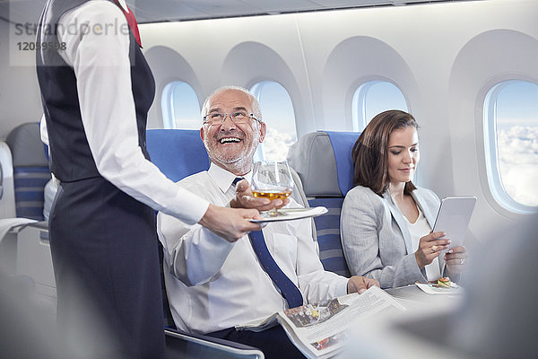 Flugbegleiter  der dem lächelnden Geschäftsmann Whiskey serviert  der in der ersten Klasse im Flugzeug sitzt.