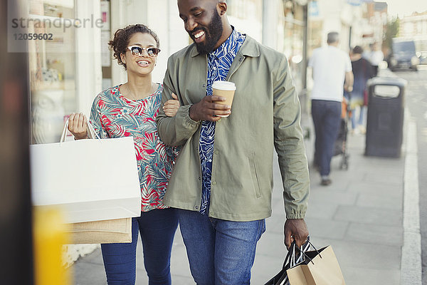 Lächelndes junges Paar mit Kaffee und Einkaufstüten  das an der Fassade entlanggeht.