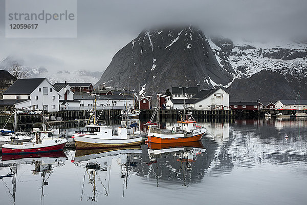 Fischerboote und Dorf am Wasser unter verschneiten  zerklüfteten Bergen  Hamnoya  Lofoten  Norwegen