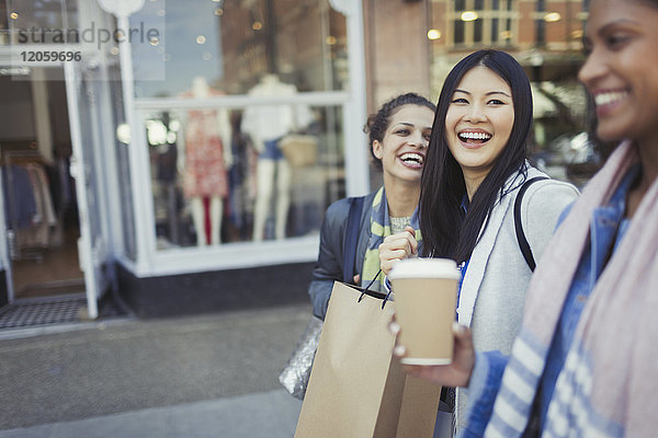 Lachende Freundinnen  die mit Kaffee und Einkaufstaschen an der Fassade entlanggehen