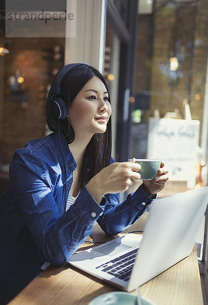 Nachdenkliche junge Frau hört Musik mit Kopfhörern und trinkt Kaffee am Laptop im Caféfenster.