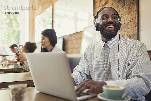 Lächelnder junger Mann mit Kopfhörer am Laptop und Kaffeetrinken am Kaffeetisch