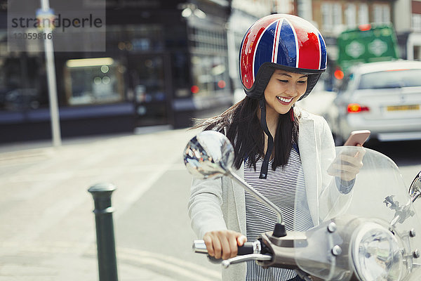 Lächelnde junge Frau beim SMSen mit Handy auf dem Motorroller  mit Helm auf der Straße.