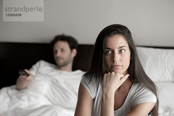 Frau sitzt im Bett  während ihr Mann fernsieht.