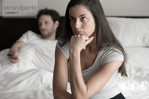 Frau sitzt im Bett  während ihr Mann fernsieht.