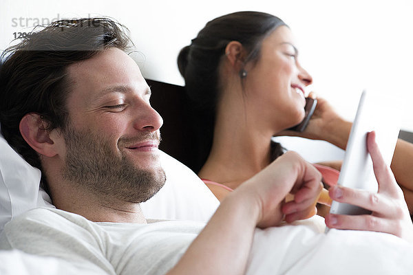Paar entspannt im Bett  Mann mit digitalem Tablett  während Frau telefoniert