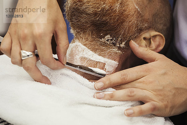 Ein Kunde sitzt auf dem Stuhl des Barbiers und lässt sich von einem Barbier mit einem Rasiermesser mit durchgeschnittener Kehle nass rasieren.