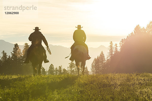 Zwei Cowboys reiten zu Pferd in einer Prärielandschaft bei Sonnenuntergang.