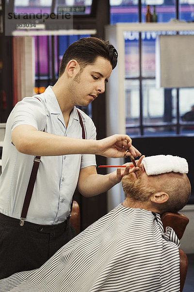 Ein Kunde  der mit einem heißen Handtuch im Gesicht auf dem Stuhl des Barbiers sitzt und dessen Bart von einem Barbier gestutzt wird.
