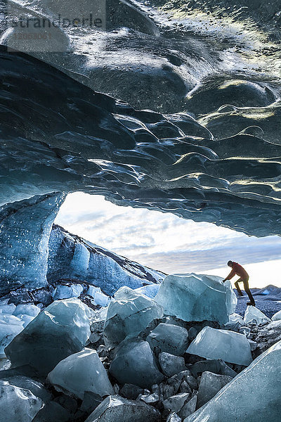 Seitenansicht einer Person  die auf Eisfelsen am Eingang einer Gletschereishöhle klettert.