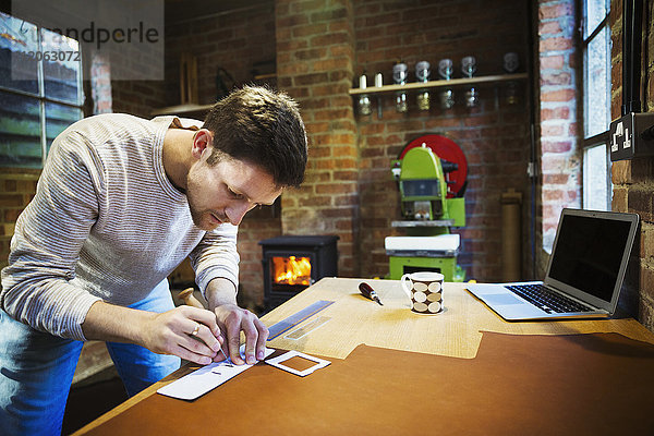 Ein Handwerker  der einen Pfriem und ein Lineal benutzt  um ein kleines Stück Leder zu messen und zu markieren. Werkbank mit einem Becher  Handwerkzeug und einem Laptop.