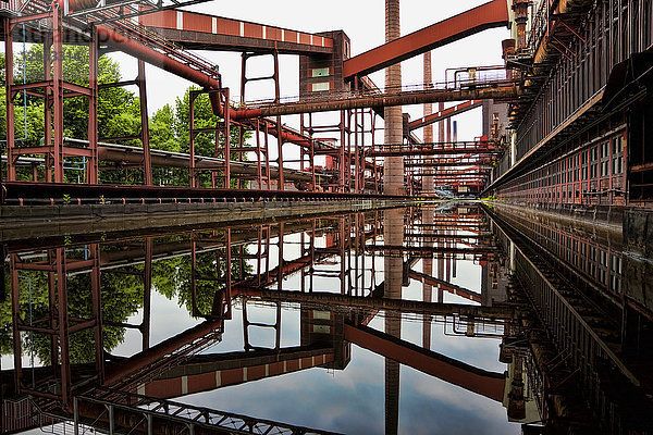 Industriegebäude mit quer verlaufenden Rohren und Schornsteinen  die sich in einem Kanal spiegeln.