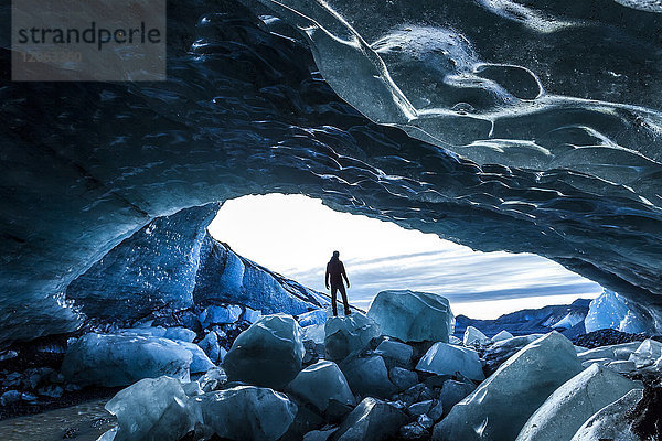 Rückansicht der Silhouette einer Person  die auf einem Eisfelsen am Eingang zu einer Gletschereishöhle steht.
