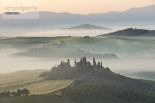 Sonnenaufgang über einer nebligen italienischen Landschaft mit sanften Hügeln und Zypressen  in der Ferne ein Bauernhaus.