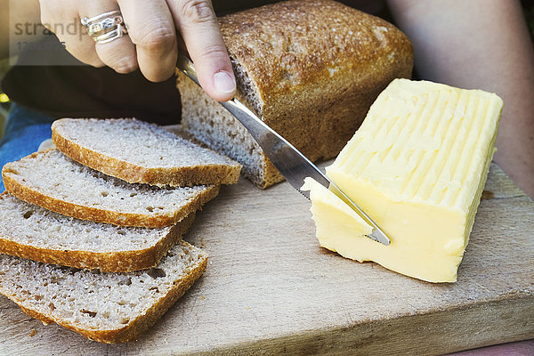 Eine Person  die mit einem Messer einen Butterblock für einen geschnittenen Brotlaib durchschneidet.
