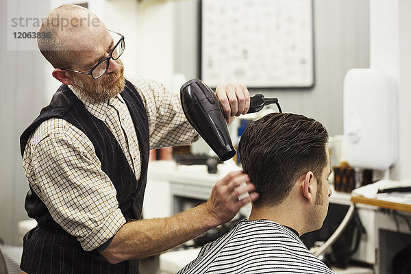 Ein Kunde  der auf dem Stuhl des Friseurs sitzt und sich von einem Friseur und Barbier die Haare fönen lässt.
