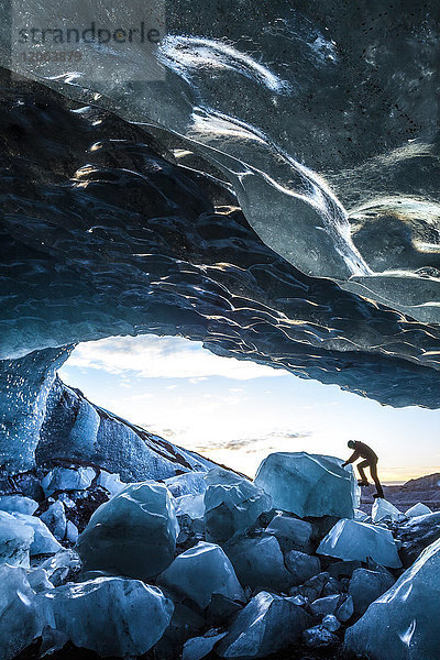 Seitenansicht einer Person  die auf Eisfelsen am Eingang einer Gletschereishöhle klettert.