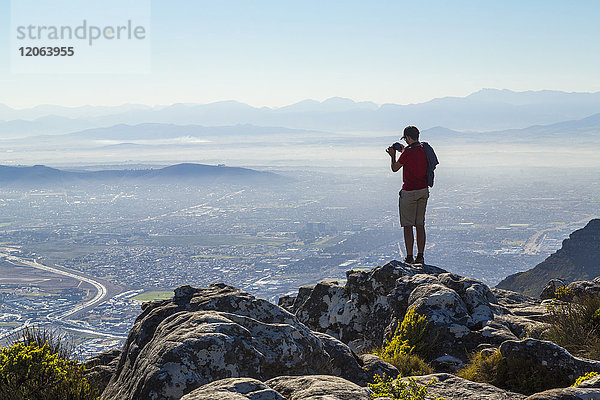 Mann steht auf einem Felsen am Rande eines Berges und fotografiert Stadtlandschaft und Gebirge in der Ferne.