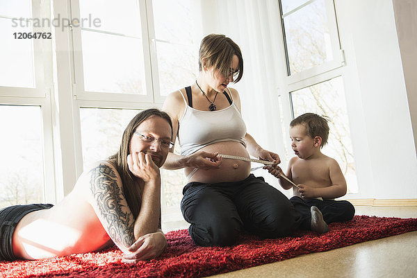 Porträt der Eltern  die mit ihrem kleinen Sohn auf einem Teppich sitzen und den schwangeren Bauch der Mutter messen
