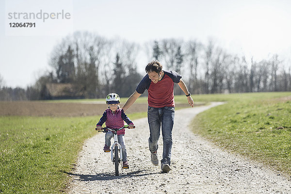 Vater hilft seiner Tochter beim Fahrradfahren auf dem Fußweg inmitten eines Feldes