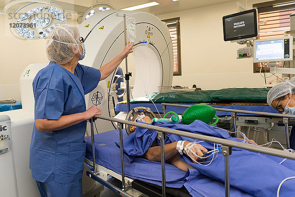 Reportage aus dem Operationssaal der stereotaktischen Neurochirurgie im Krankenhaus Pasteur 2  Nizza  Frankreich. Behandlung der Parkinson-Krankheit durch tiefe Hirnstimulation. Bei dieser Technik werden Elektroden in das Gehirn implantiert  die die abnorme elektrische Aktivität des Gehirns modulieren. Der Raum ist mit einem O-Arm-Scanner für die OP-Vorbereitung und dem NeuroMate  einem stereotaktischen Roboter  ausgestattet. Der Roboter ermöglicht es den Chirurgen  ihre Operationstechnik zu optimieren und zuverlässiger zu gestalten  und der O-Arm ermöglicht 3D-Bilder in Echtzeit  um die Präzision  Sicherheit und Effizienz der Operation zu verbessern. Einrichten des Patienten.