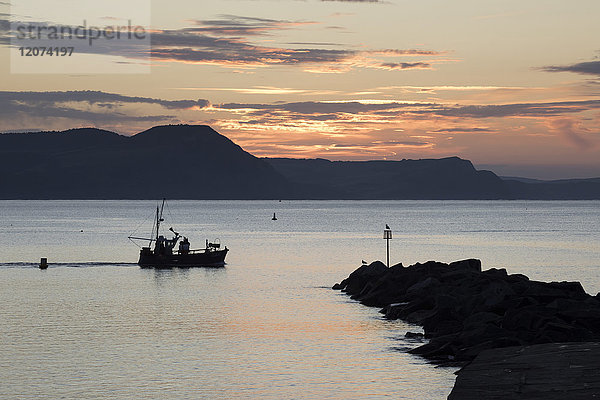 Fischerboot  das bei Sonnenaufgang an The Cobb vorbeifährt  mit Golden Cap und Jurassic Coast  UNESCO-Weltkulturerbe  Lyme Regis  Dorset  England  Vereinigtes Königreich  Europa